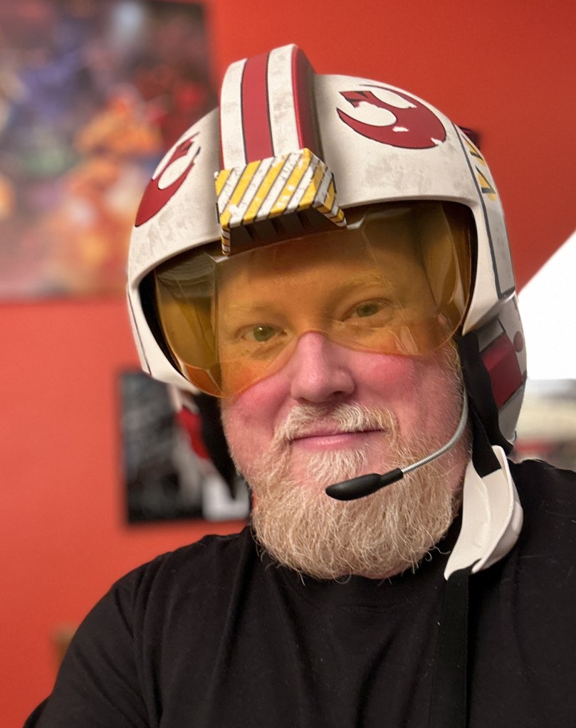 a picture of the author with a blond beard, wearing an x-wing flight helmet against an orange background